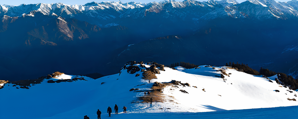 Trekkers climbing snow-covered Kedarkantha peak with Himalayan range glowing in golden sunrise.