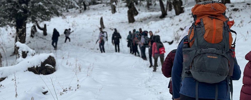 Trekkers walking through snow-covered Kuari Pass trail with panoramic views of Nanda Devi and surrounding Himalayan peaks.